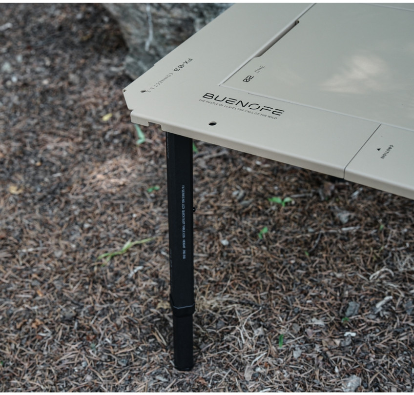 BUENOFE IGT Quick Slot Table Leg shown in an outdoor close-up where a black leg meets the corner of a tan IGT tabletop engraved BUENOFE FX-L03 Connect 1.5, detailing the twist-lock connection point over a forest floor.