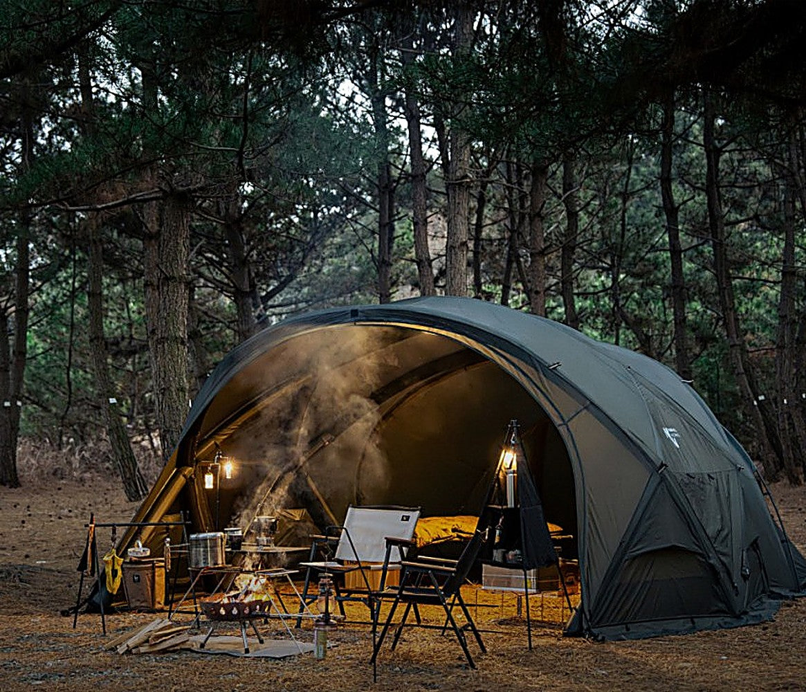 Evening campsite with the Shelter GM lit from within, campfire smoke drifting past chairs in a pine forest