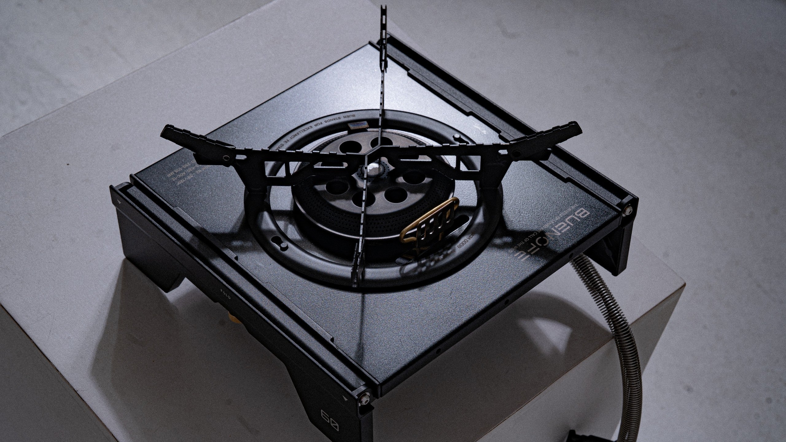 BUENOFE Stove Windscreen installed on the BX-09 Table Burner in an IGT frame, photographed from above on a grey concrete surface in moody directional light that emphasizes the matte black ring and cross pot supports.