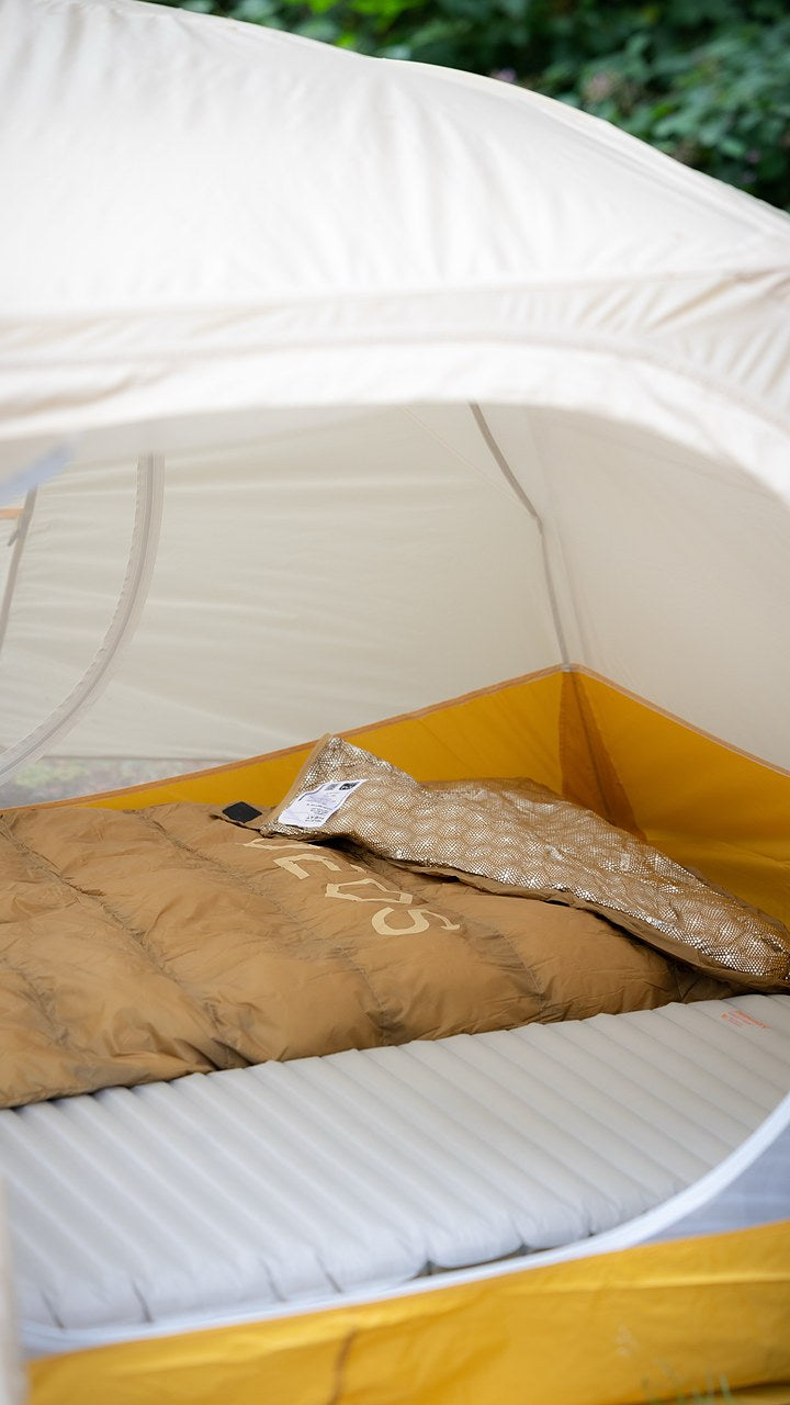 Field-tester photo — tan DEOS camp comforter laid out on a mat inside a tent