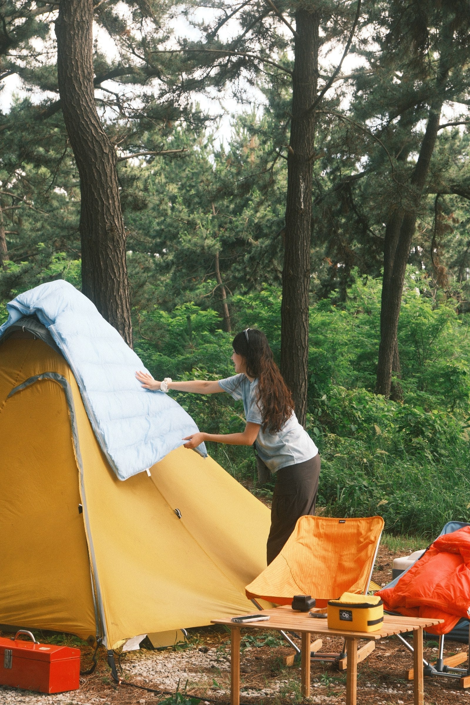 Camper holding a sky blue DEOS camp comforter open beside a tent in the forest