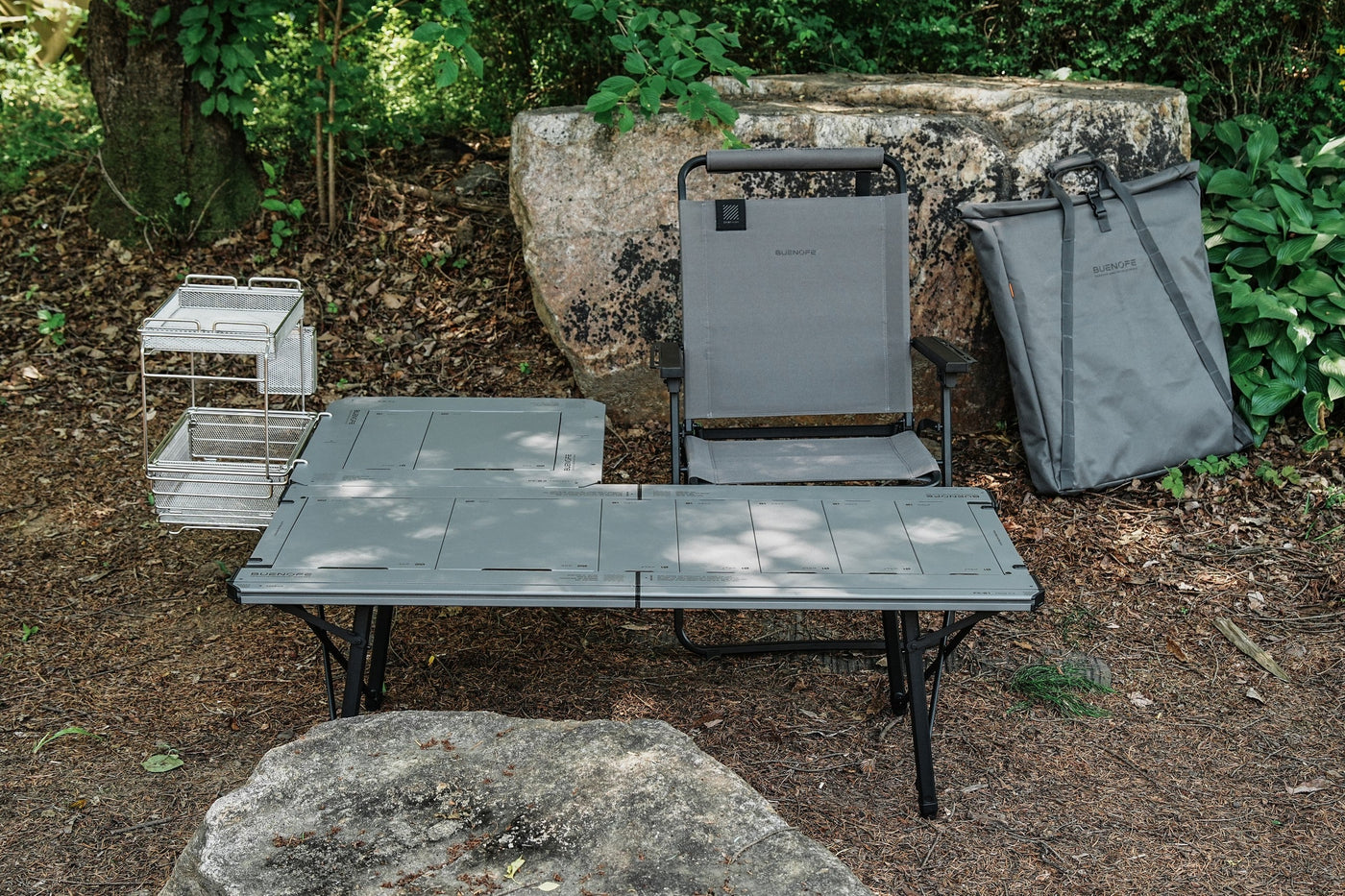 BUENOFE IGT Connect Table 1.5 graphite-gray table set up outdoors beside a matching padded carry bag, a camp chair and a stainless wire rack against mossy rocks.