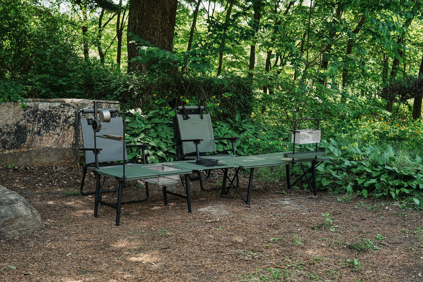 Wide forest scene of a long olive BUENOFE IGT Connect Table 1.5 camp table flanked by two camp chairs beside a large boulder, styled as a group lounge and cooking setup.