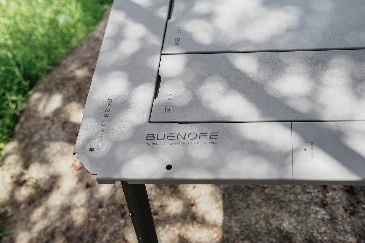 Detail of the laser-engraved surface of a silver-gray BUENOFE IGT Connect Table 1.5, showing the FX-03 CONNECT 1.5 model marking and BUENOFE wordmark with the tagline The Rustle of Leaves The Call of the Wild under dappled shade.