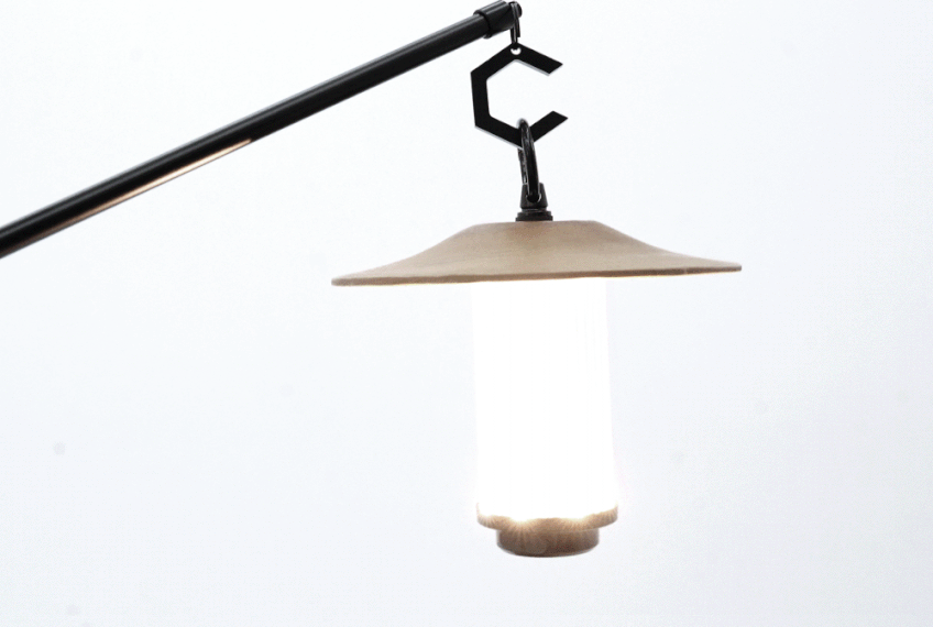 BUENOFE IGT Lantern Hanger holding a warmly glowing lantern from its black C-hook at the end of the boom arm, the lit lantern casting soft light against a pale background to suggest an evening campsite mood.