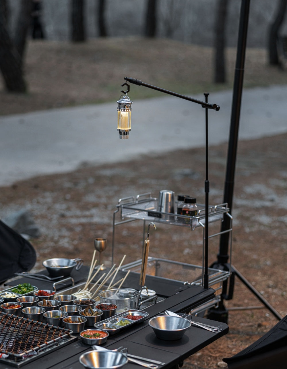 BUENOFE IGT Lantern Hanger holding a glowing lantern over an evening camp cooking setup with skewers and stainless bowls on a black IGT table.