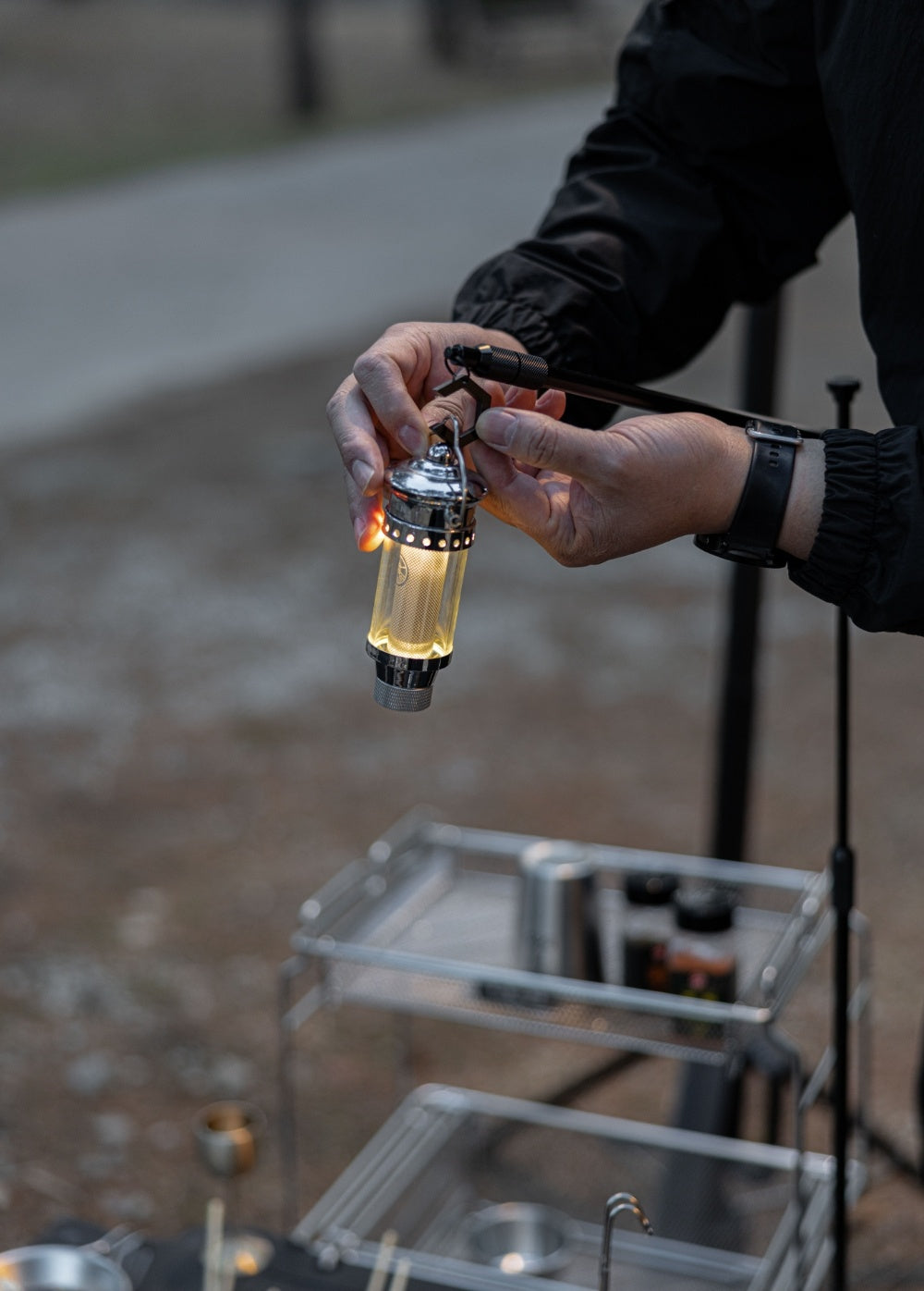 BUENOFE IGT Lantern Hanger at a dusk campsite: hands clip a small lit lantern onto the hanger arm above an outdoor cooking table.