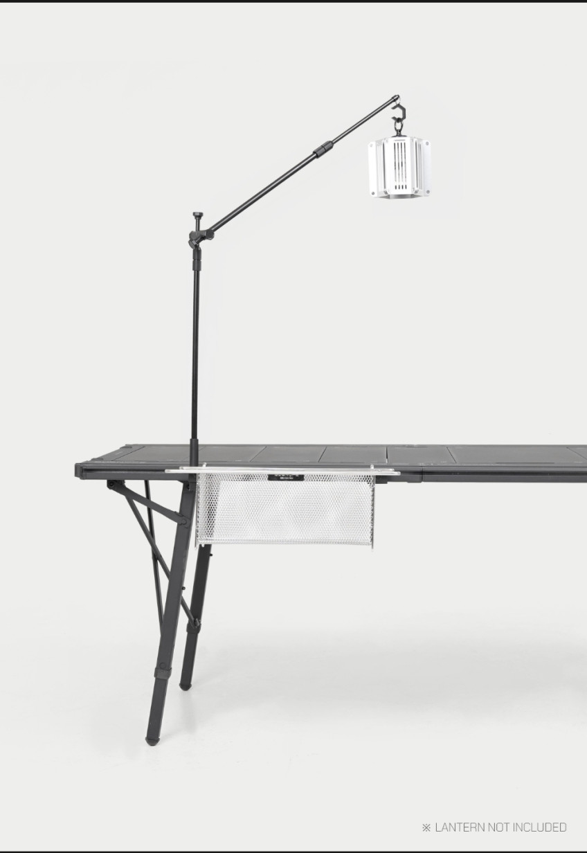 BUENOFE IGT Lantern Hanger in a side profile view mounted to a camping table, the boom arm reaching across the tabletop with a lantern and a mesh storage pocket slung under the rail; an on-image note reads that the lantern is not included.