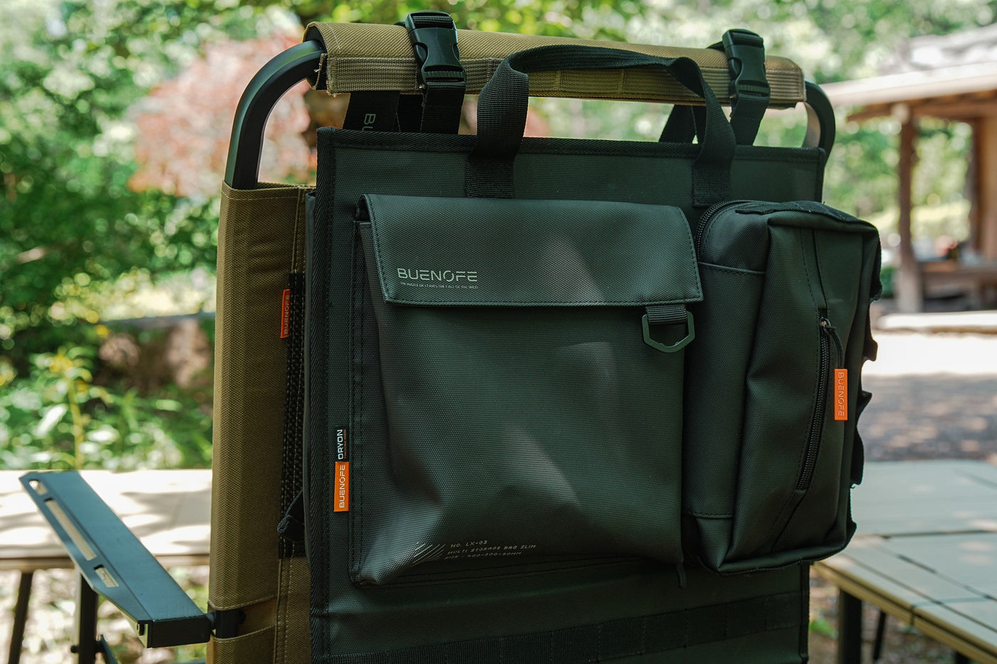 BUENOFE Multi Storage Bag Slim close-up while mounted on a camp chair in warm daylight, showing the front flap pocket with the BUENOFE logo and embossed model text, the D-ring, the side tissue pouch and the orange zipper pulls.