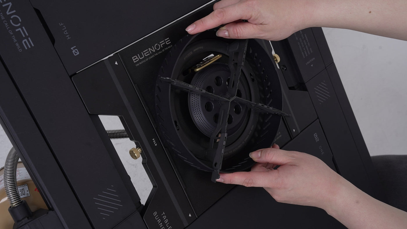Hands lowering the BUENOFE Stove Windscreen ring onto the BX-09 Table Burner head seated in an IGT table, shown from the side to illustrate the tool-free drop-on installation of the wind shield.