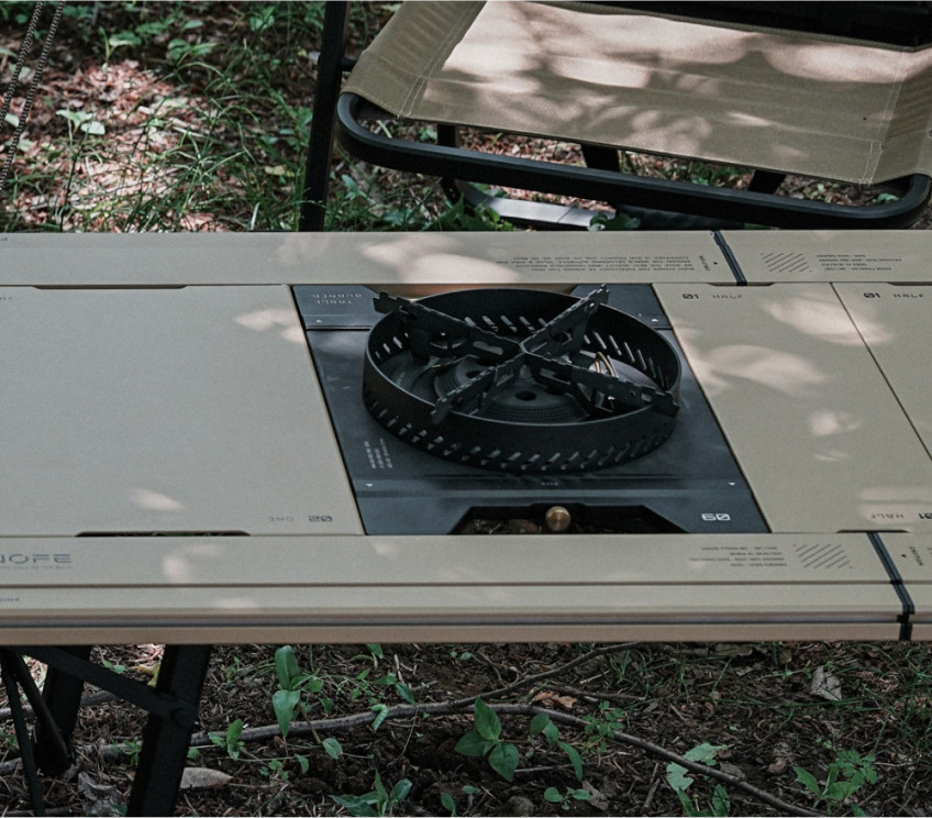 BUENOFE Stove Windscreen ringing the BX-09 Table Burner on an outdoor IGT table, viewed from above in dappled forest light with foliage and camp gear softly blurred in the background.