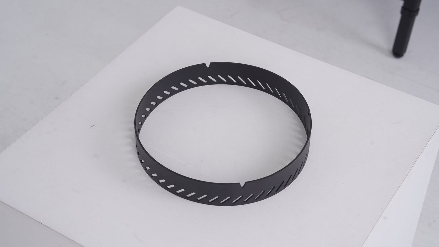 BUENOFE Stove Windscreen shown on its own as a matte black stainless steel ring resting on a white surface in a three-quarter view, highlighting the circular band and the row of angled diagonal air vents cut around its wall.