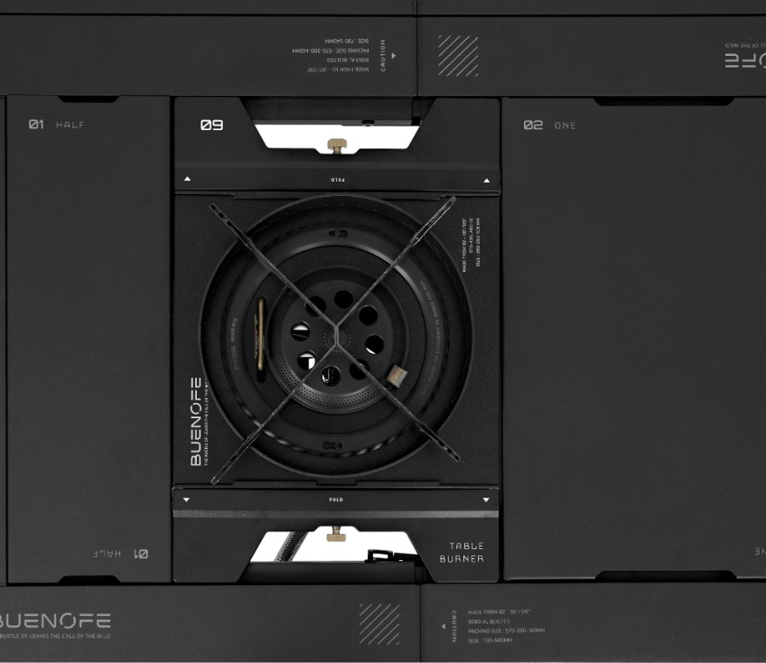 Overhead technical studio view of the BUENOFE Stove Windscreen around the BX-09 Table Burner head, showing the vented ring, cross pot supports and the burner ports in crisp detail against a dark IGT table with fine English spec markings.