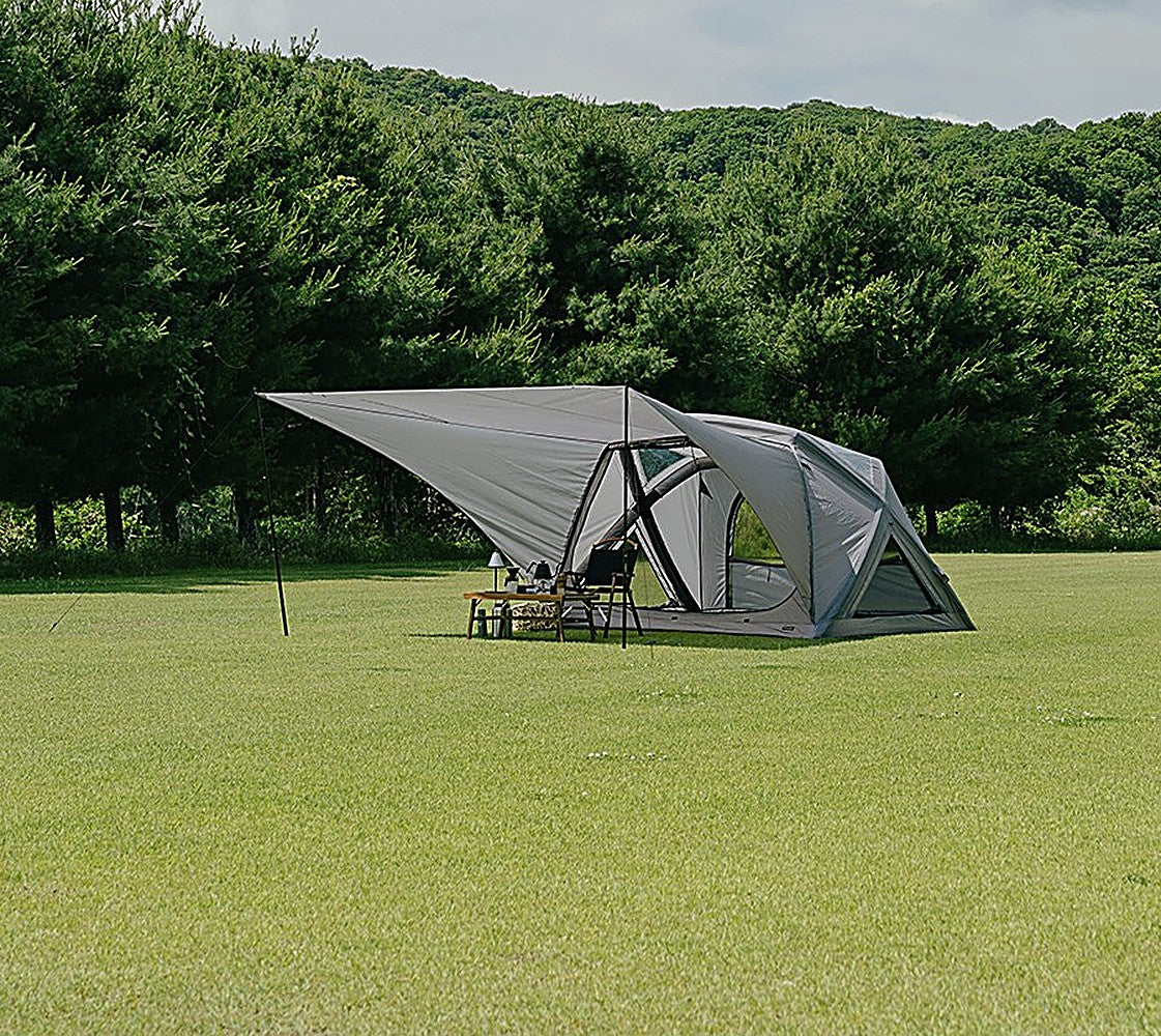 Distant view of the Extension Sun Shade and GEO AIR4 tent set up on an open green field ringed by forest