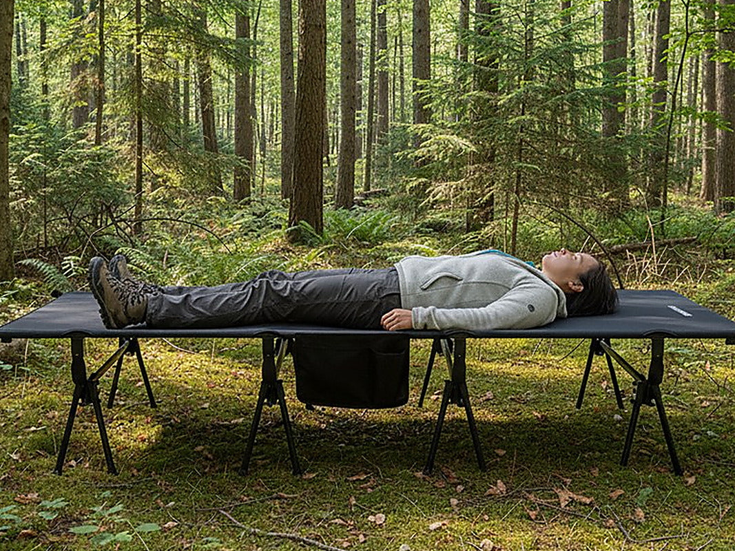 A camper lying on the Wide Cot 80 in a forest clearing, arms resting on the cot.